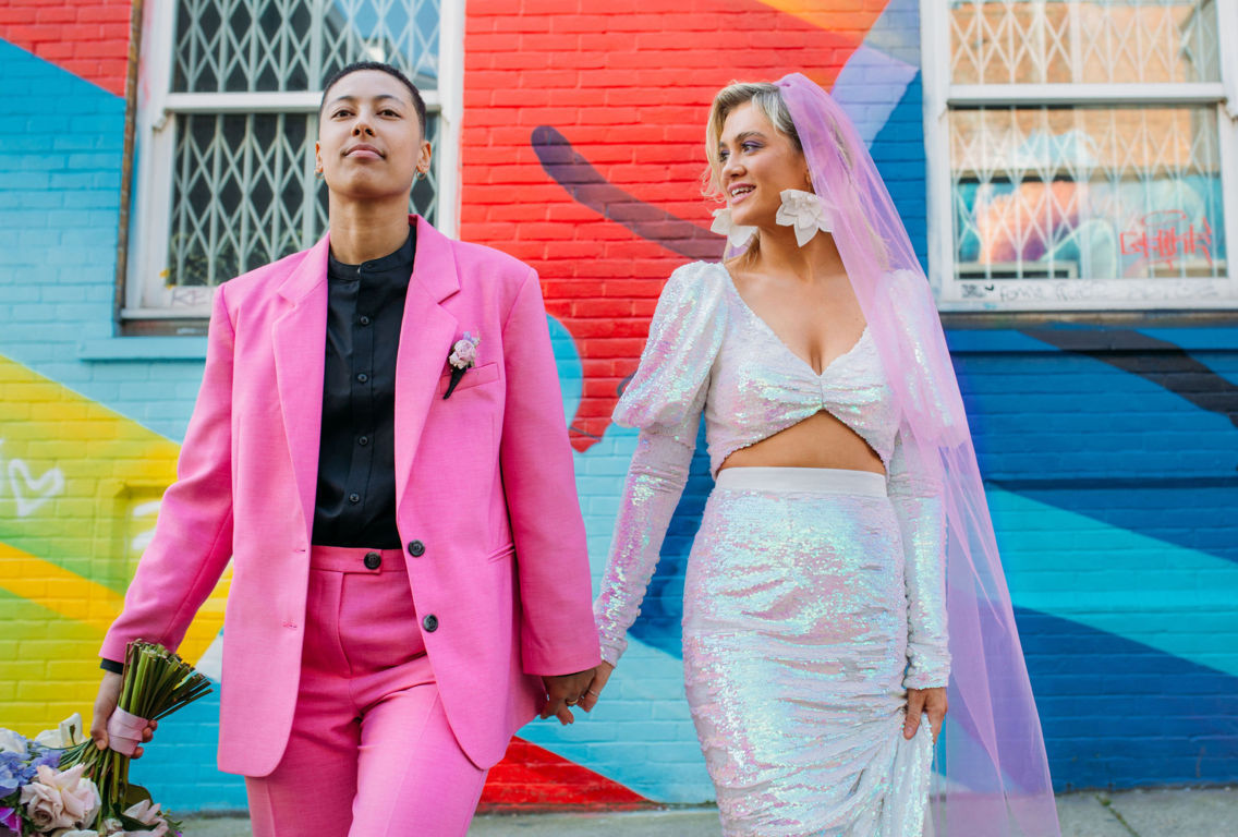 a bride with statement earrings, an iridescent, sequinned 2 piece wedding dress and loose tousled hair and a groom with a grade 1 hair style wearing a bright pink suit, black shire and gold jewellery 
