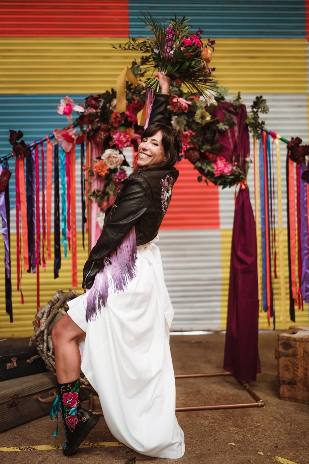 UK wedding inspiration - smiling bride lifts up her skirt to show off bespoke hand painted doc marten boots whilst holding a bouquet up to the sky in front of a party streamer background. Her leather jacket has bright pink tassels