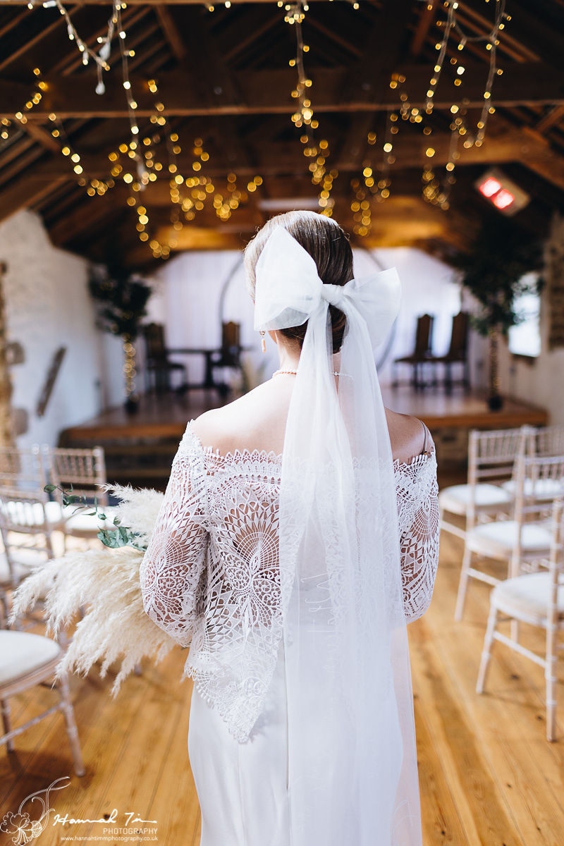 portrait image of the bride walking down the aisle surrounded with soft wash chiavari chairs in a boho lace dress, white hair bow veil and a clean up do at Chypraze Barn