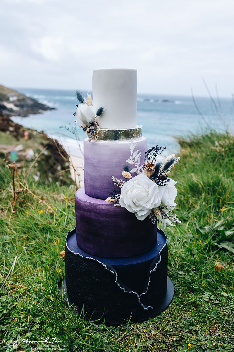 three tier wedding cake in ombre shading starting with a deep purple at the base and ending with white at the top adorned with dried flowers on the right hand side, and the top positioned outside Chypraze Barn overlooking the Cornish Coast